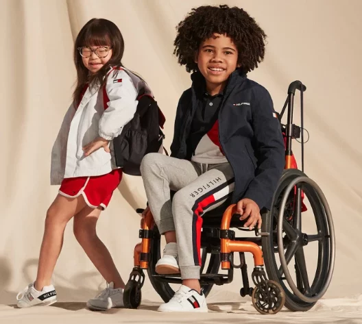 A black boy in wheelchair and a girl with glasses standing next to her, both wearing Tommy Hilfiger