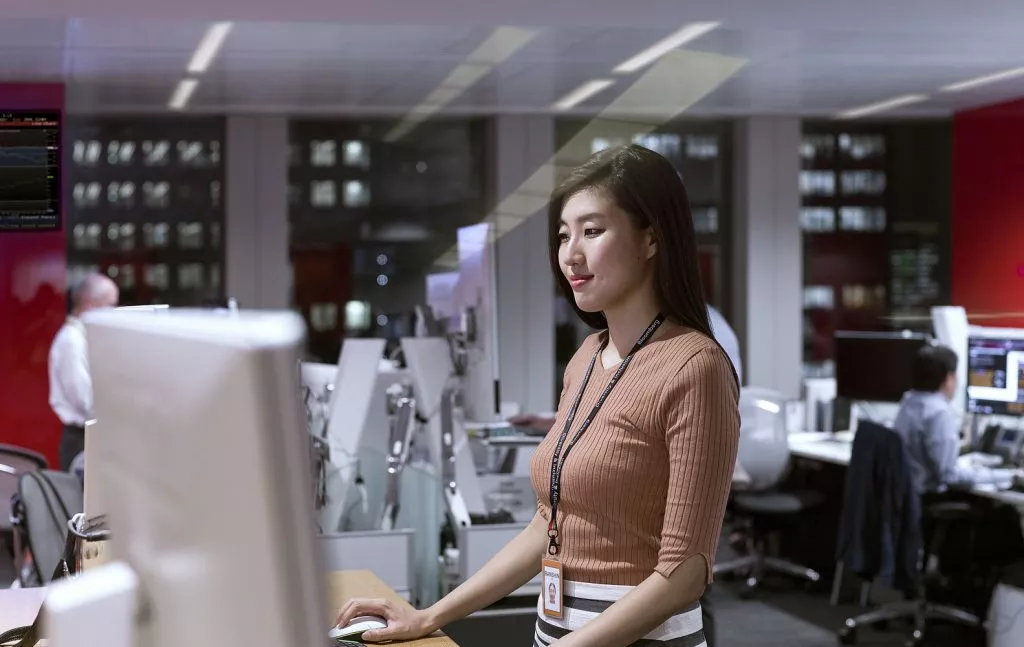 Asian woman standing in front of a desktop in an office, smiling and researching something