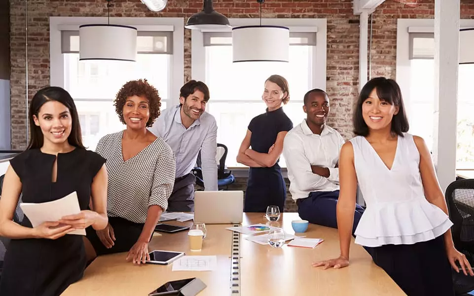 A diverse group of young men and women gather at an office conference table, smiling