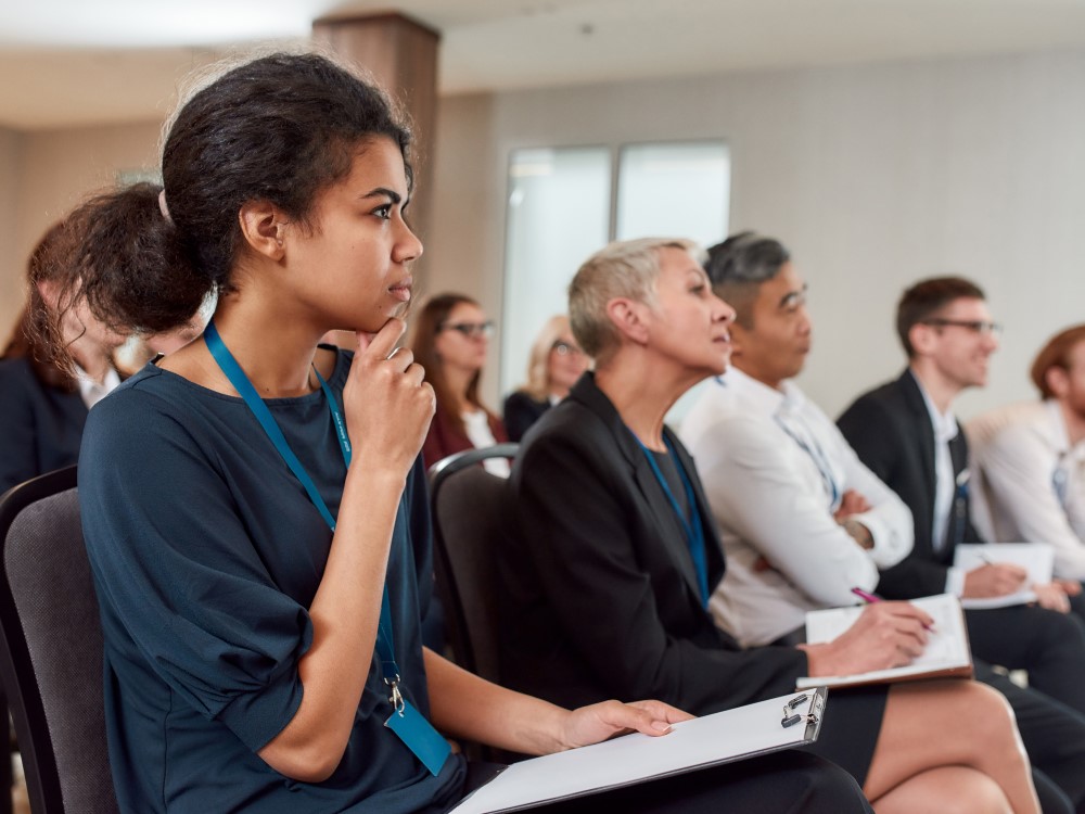 A black woman and other audience facing forward, listening to presentation, and taking notes