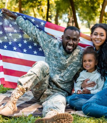 African American military man sitting on the lawn with a flag, smiling with his wife and children, a soccer ball and a basket